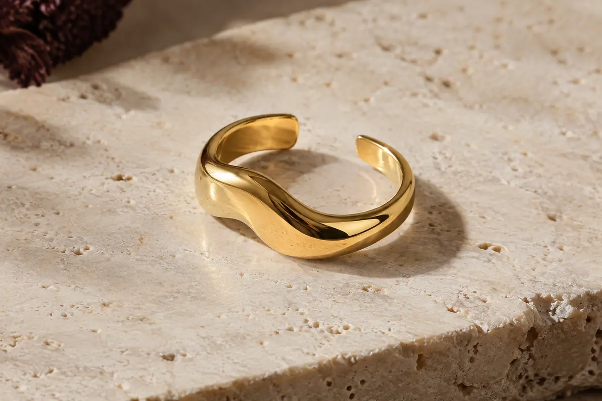 Bague ouverte dorée au profil ondulé, posée sur une surface de travertin clair.